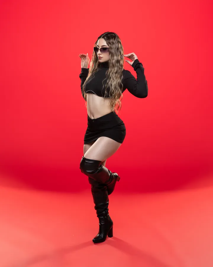 Miss Lu de cuerpo entero sobre fondo rojo, con gafas de sol y botas altas.