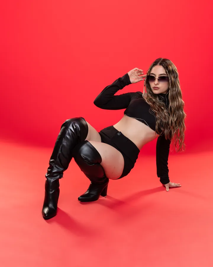 Miss Lu apoyada sobre fondo rojo, con gafas de sol y botas altas.